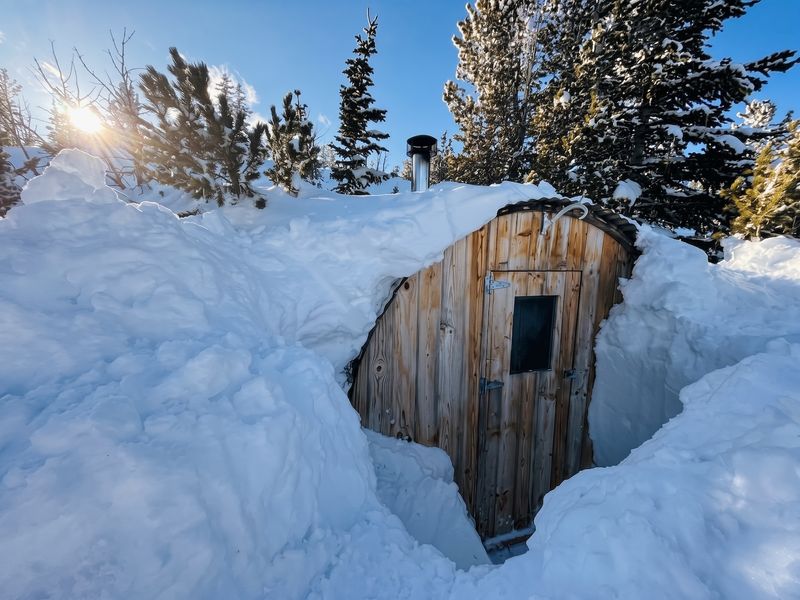 Big Belt Hut sauna buried in deep snow beneath sunlit trees